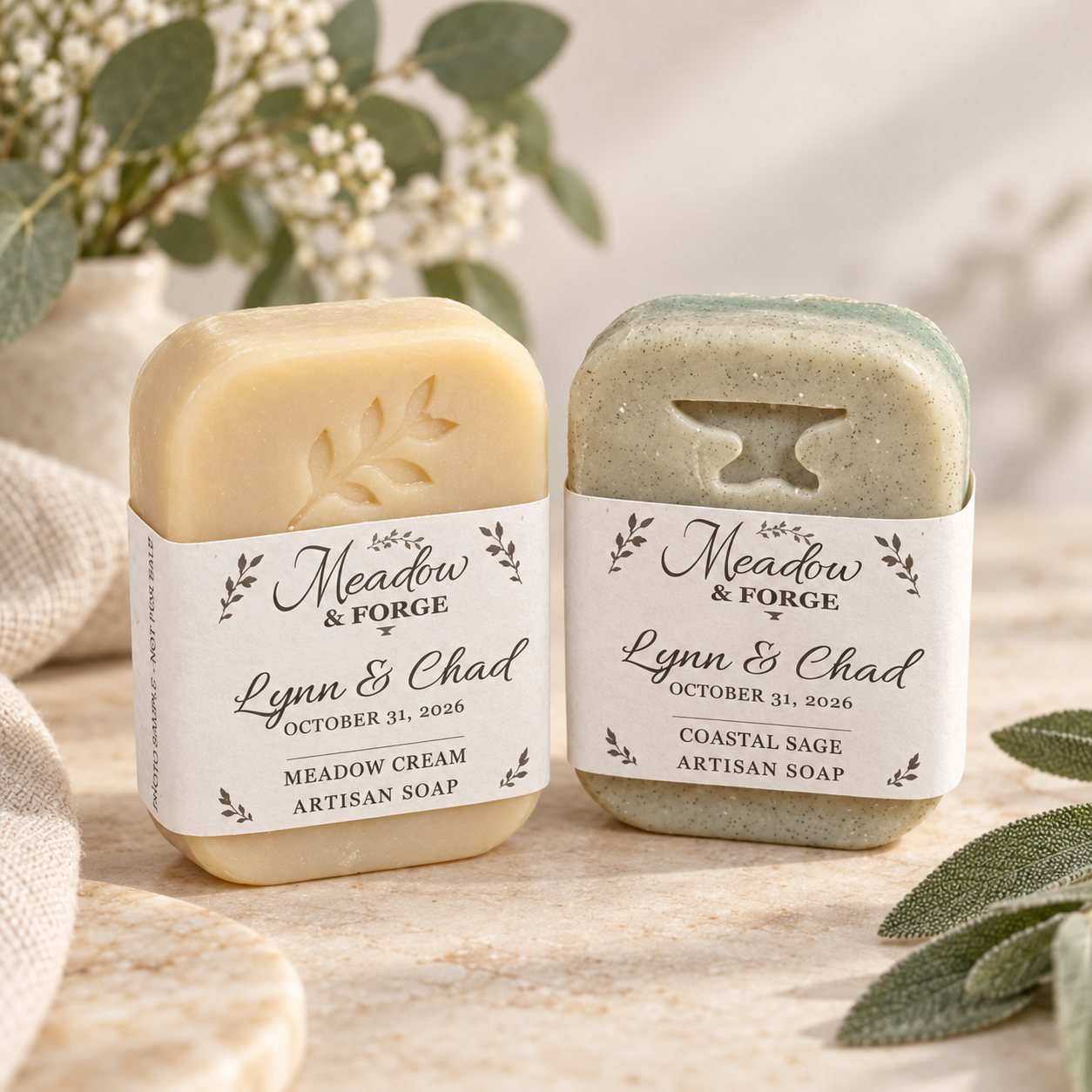 A cream Meadow Cream bar stamped with a leaf and a sage Coastal Sage bar stamped with an anvil, each in a white Meadow & Forge band reading Lynn & Chad, October 31, 2026, on pale stone with linen and greenery.